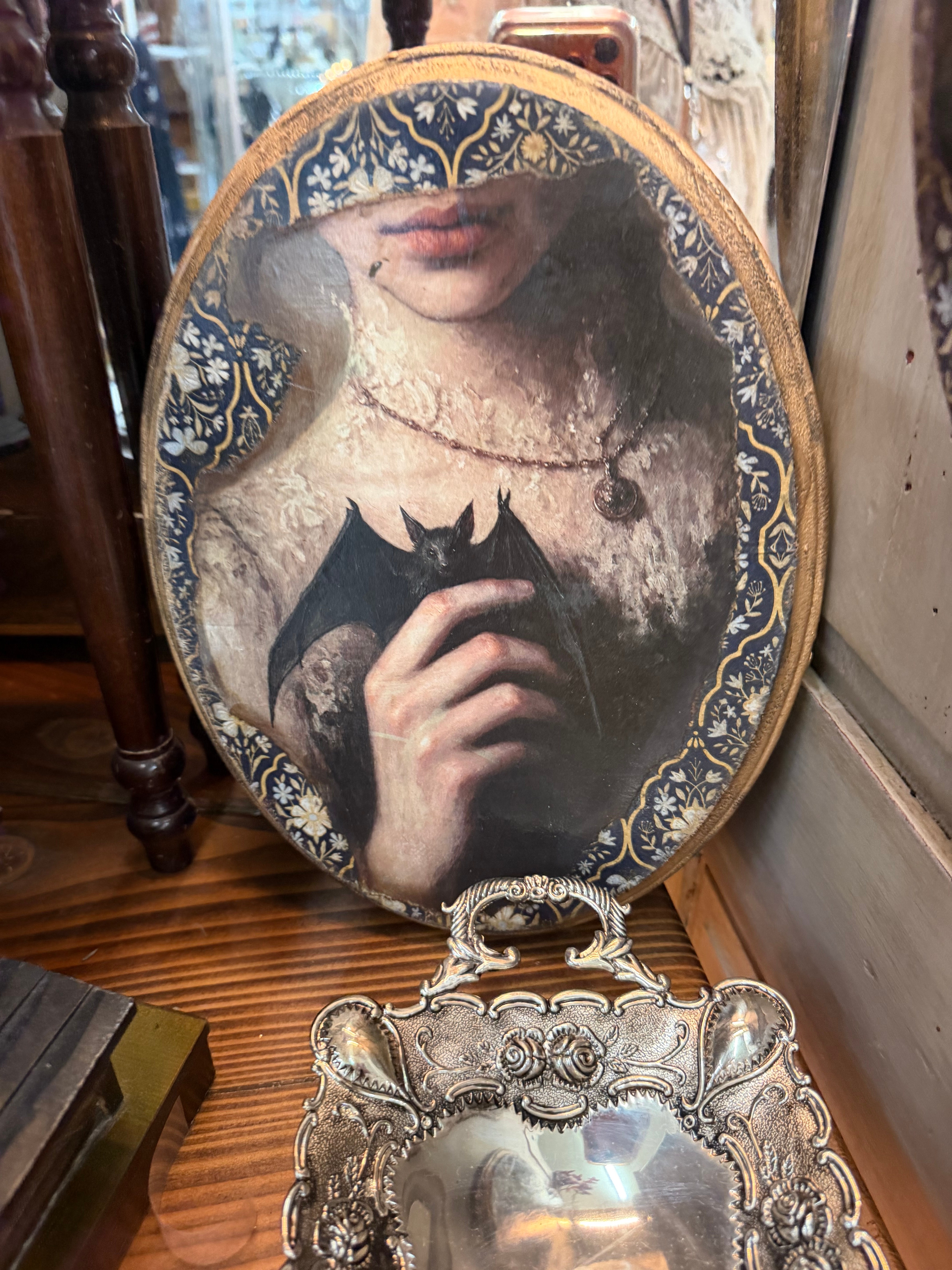 An oval decoupage art plaque of a Victorian woman holding a bat, bordered in blue and gold florals