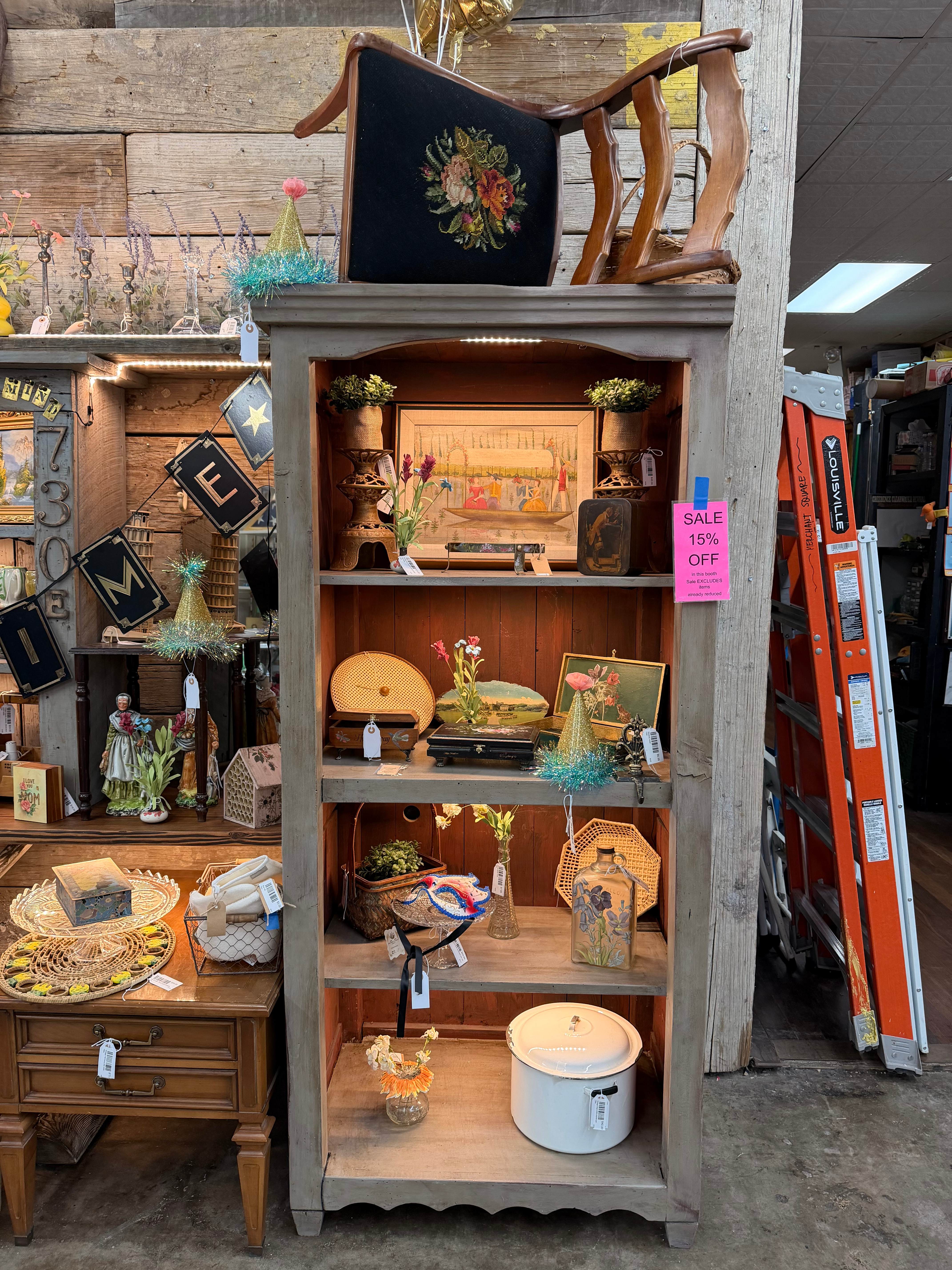 A tall gray hutch styled with a needlepoint chair on top, framed folk art, baskets, an enamel pot, party hats, and the 730E booth letters
