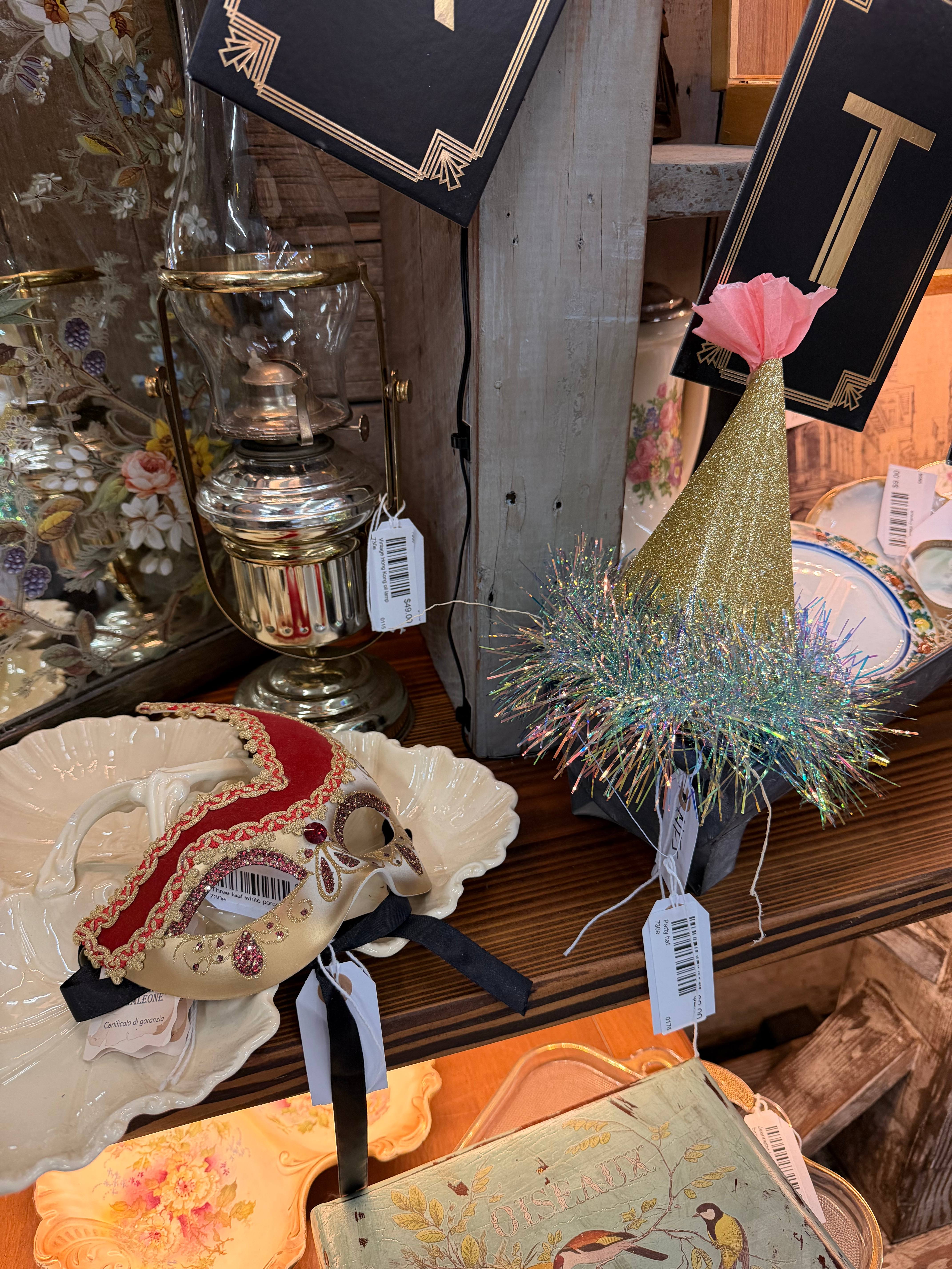 A close-up vignette of a brass oil lamp, a gold tinsel party hat, and an ornate red and gold Venetian mask on a white scalloped plate