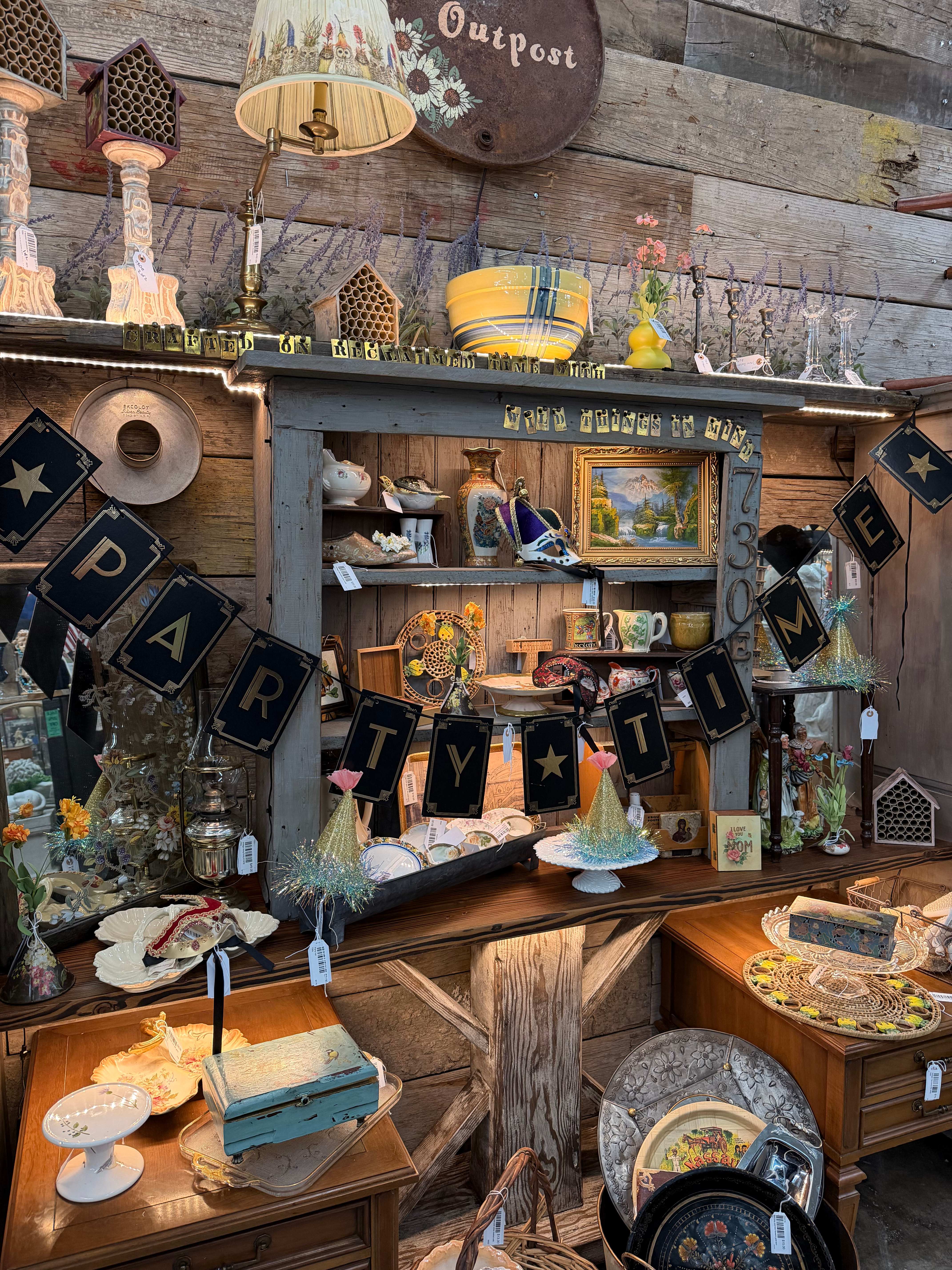 A styled Party Time vignette with the sunflower Outpost sign, lavender sprigs, a yellow striped bowl, glittery party hats, masquerade masks, and vintage cake stands