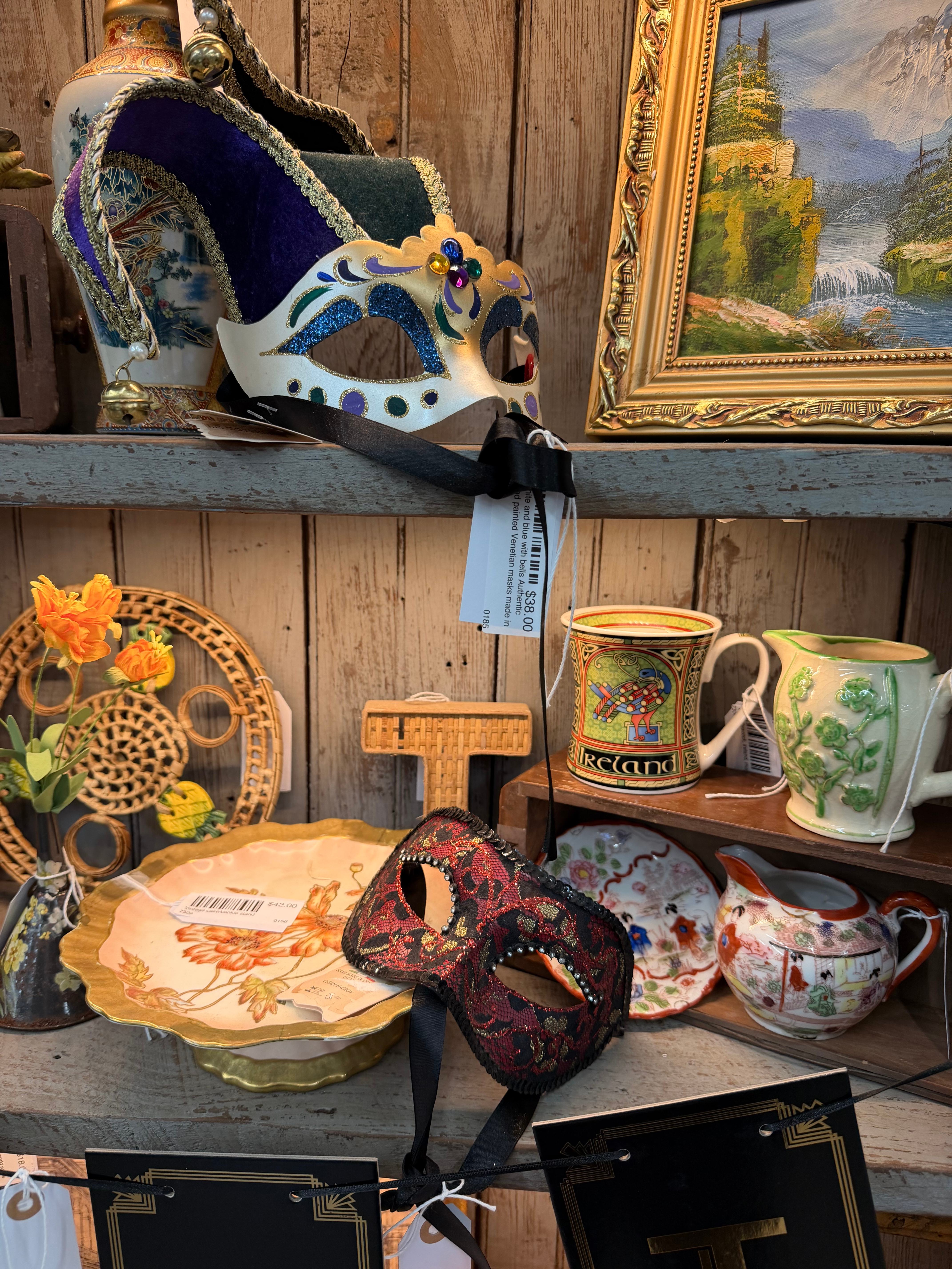 A shelf grouping of Venetian masquerade masks, an Ireland mug, a green shamrock pitcher, floral plates, and a gold-framed waterfall painting